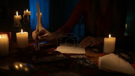 Téléchargez les photos : Gros plan d'une femme assise sur la table avec beaucoup de bougies autour d'elle. Elle écrit quelque chose avec un stylo plume sur le vieux papier. Concept esthétique magique. - en image libre de droit