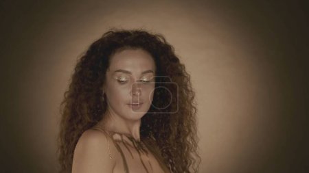 Photo for Close up shot of a young attractive woman shyly looking to the side, golden makeup and accessories. Golden drips. Brown gradient background. Beauty and cosmetics advertisement. - Royalty Free Image