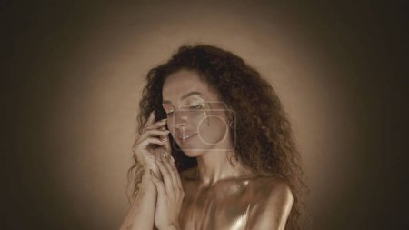 Photo for Close up shot of a young attractive woman looking to the side and touching her face with fingers, golden makeup and accessories. Golden drips. Brown background. Beauty and cosmetics advertisement. - Royalty Free Image