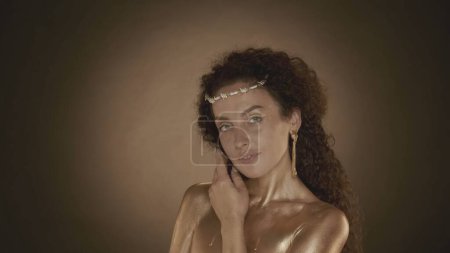 Photo for Close up shot of a young attractive woman looking at the camera, touching her neck. Golden makeup, earrings and crown of thorns. Golden paint. Brown background. Beauty and cosmetics advertisement. - Royalty Free Image