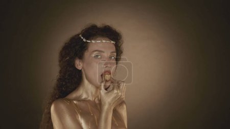 Photo for Close up shot of a young attractive woman looking at the camera, eating a candy. Golden makeup, earrings and crown of thorns. Golden paint. Brown background. Beauty and cosmetics advertisement. - Royalty Free Image