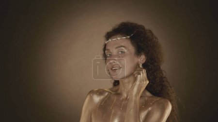 Photo for Close up shot of a young and attractive woman looking at the camera smiling touching her ear, golden makeup and accessories. Golden drips. Brown gradient background. Beauty and cosmetics advertisement - Royalty Free Image