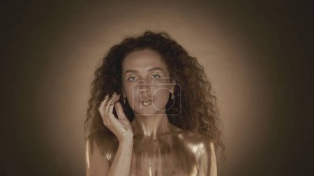 Photo for Close up shot of a young attractive woman looking to the side and touching her face with fingers, golden makeup and accessories. Golden drips. Brown background. Beauty and cosmetics advertisement. - Royalty Free Image