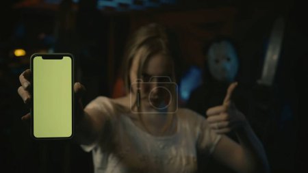 Photo for Girl victim of the horror maniac, standing and holding a smartphone, showing thumbs up at the camera. Man in hockey mask standing behind. Advertising area, workspace mockup. - Royalty Free Image