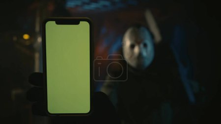 Photo for Maniac in white hockey mask holding showing a smartphone at the camera, standing inside the warehouse. Focus on the screen. Advertising area, workspace mockup. - Royalty Free Image