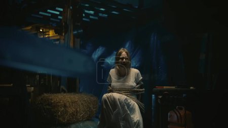 Photo for Horror movie scene. Closeup shot of the chainsaw. Female victim of the maniac in a white dress sitting on a chair tied with a rope at the background. - Royalty Free Image