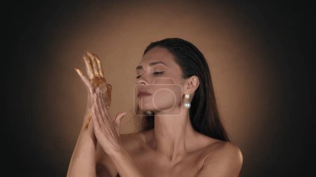 Téléchargez les photos : Gros plan d'un modèle féminin avec des boucles d'oreilles en perles regardant la peinture dorée couler de sa main et la toucher. Concept de publicité beauté. - en image libre de droit