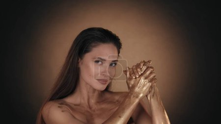 Photo for Portrait of an attractive woman. Close up shot of female model with shiny makeup applying golden liquid paint on her hands skin, looking at the camera. Beauty advertisement concept. - Royalty Free Image