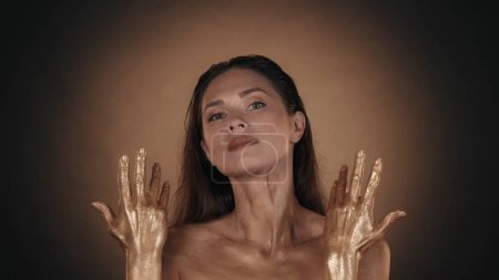 Téléchargez les photos : Portrait d'une femme brune au maquillage éclatant. Gros plan du modèle féminin montrant de la peinture liquide dorée sur ses mains, en regardant la caméra. Concept de publicité beauté. - en image libre de droit