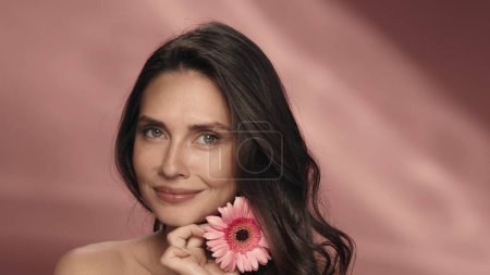 Téléchargez les photos : Une femme touche une fleur de gerbera à sa peau de visage parfaite. Portrait d'une femme avec une fleur de gerbera en studio sur fond rose en gros plan. Produits cosmétiques à l'extrait, parfum de - en image libre de droit