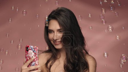 Téléchargez les photos : Une femme utilisant un smartphone prend un selfie entouré de bulles de savon. Seminude femme avec les cheveux ondulés lâches dans le studio sur un fond rose. Le concept de beauté, cosmétologie, soins de la peau, spa - en image libre de droit
