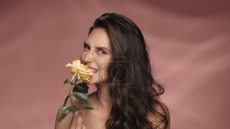 Téléchargez les photos : Une femme séminude tient une rose jaune sur une longue tige. Une femme attrape un pétale de rose avec ses dents dans un studio sur un fond rose. Source d'inspiration printemps et été. Le concept de parfumerie, cosmétique - en image libre de droit