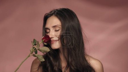 Téléchargez les photos : Une femme séminude tient une rose rouge et inhale son parfum. Une femme dans un studio sur fond rose de près. Source d'inspiration printemps et été. Le concept de parfumerie, cosmétique, soin de la peau - en image libre de droit