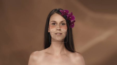 Téléchargez les photos : Portrait d'une femme séminude avec des fleurs d'orchidée dans ses cheveux et des taches d'hydrogel rose sous ses yeux. Une femme sur fond brun dans le studio. Le concept de beauté, de cosmétologie - en image libre de droit