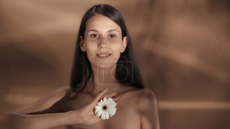 Foto de Una mujer sostiene una flor de gerberas blancas. Una joven seminude mujer en un estudio sobre un fondo marrón con reflejos. Belleza femenina natural - Imagen libre de derechos