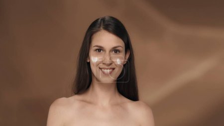 Téléchargez les photos : Une femme prend soin de sa peau, applique de la crème sur son visage. Portrait d'une jeune séminude en studio sur fond brun. Le concept de beauté, de cosmétologie, de soins - en image libre de droit