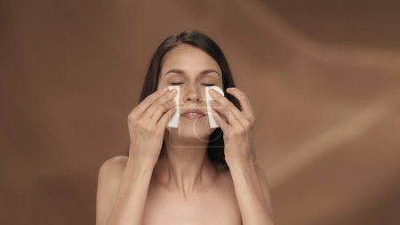 Téléchargez les photos : Une femme utilise des tampons de coton pour nettoyer son visage. Portrait d'une jeune séminude en studio sur fond brun. Le concept de beauté, de cosmétologie, de soins - en image libre de droit