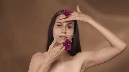 Téléchargez les photos : Une femme avec des fleurs d'orchidée dans ses mains touche son visage, appréciant la peau lisse et douce. Portrait d'une séminude en studio sur fond brun en gros plan. Le concept de beauté, de cosmétologie - en image libre de droit