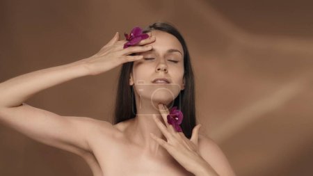 Téléchargez les photos : Une femme avec des fleurs d'orchidée dans ses mains touche son visage, appréciant la peau lisse et douce. Portrait d'une séminude en studio sur fond brun en gros plan. Le concept de beauté, de cosmétologie - en image libre de droit