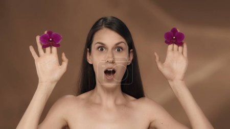 Foto de Retrato de una mujer con una expresión sorprendida, sosteniendo brotes de orquídea en sus manos. Una mujer en el estudio sobre un fondo marrón de cerca. Presentación de su producto - Imagen libre de derechos