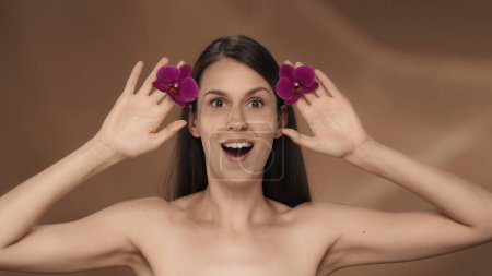 Foto de Retrato de una mujer con una expresión sorprendida, sosteniendo brotes de orquídea en sus manos. Una mujer en el estudio sobre un fondo marrón de cerca. Presentación de su producto - Imagen libre de derechos