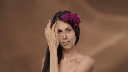 Téléchargez les photos : Portrait d'une séminude avec des fleurs d'orchidée dans les cheveux sur fond brun en studio. Le concept de beauté, de cosmétologie, de traitement et de soins - en image libre de droit