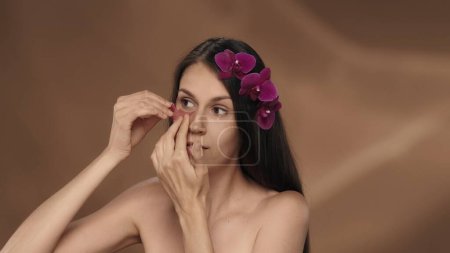 Téléchargez les photos : Femme appliquant, lissant les taches d'hydrogel rose sous ses yeux. Portrait d'une séminude avec des fleurs d'orchidée dans ses cheveux en studio sur fond brun. Le concept de beauté, de cosmétologie - en image libre de droit