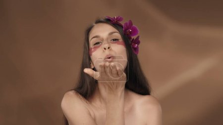 Photo for A woman with hydrogel patches under her eyes blows a kiss. Seminude woman with orchid flowers in her hair in the studio on a brown background. Beauty, cosmetology and care - Royalty Free Image