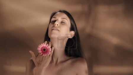 Téléchargez les photos : Une femme touche sa main avec une fleur de gerbera rose sur la peau de son cou. Une jeune séminude démontre sa peau parfaite dans un studio sur fond brun. Le concept de beauté, de cosmétologie - en image libre de droit