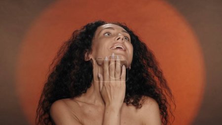 Photo for A woman enjoys the softness and silkiness of her skin, touches her neck. Portrait of a seminude curly woman in the studio on an orange background with circular light. The concept of beauty - Royalty Free Image