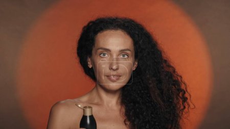 Téléchargez les photos : Portrait d'une femme avec une bouteille noire de produit cosmétique, lotion, crème, baume. Seminude femme bouclée en studio sur fond orange avec lumière circulaire. Le concept de beauté, cosmétologie, soin de la peau - en image libre de droit