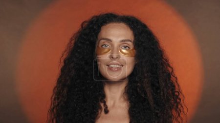 Photo for Longhaired curly brunette woman performs cosmetic procedures. Portrait of a seminude woman with golden patches under her eyes in the studio on an orange background with a circular light close up. The - Royalty Free Image