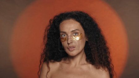 Téléchargez les photos : Longue femme brune bouclée effectue des procédures cosmétiques. Portrait d'une séminude avec des taches dorées sous les yeux en studio sur fond orange avec une lumière circulaire en gros plan. L ' - en image libre de droit