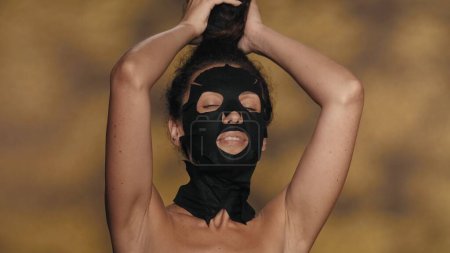 Téléchargez les photos : Une femme avec un masque en drap cosmétique noir et un chignon sur la tête. Séminude femme effectuant des procédures cosmétiques dans le studio sur un fond jaune avec des faits saillants. Le concept de beauté - en image libre de droit