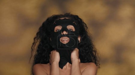 Téléchargez les photos : Une femme avec un masque en drap cosmétique noir. Séminude femme effectuant des procédures cosmétiques dans le studio sur un fond jaune avec des faits saillants. Le concept de beauté, de cosmétologie, de soins - en image libre de droit