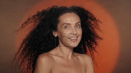 Téléchargez les photos : Seminude femme sur un fond orange avec une lumière circulaire. De longs cheveux bouclés et épais d'une femme flottant dans le vent. Le concept de beauté, coiffure, soins - en image libre de droit