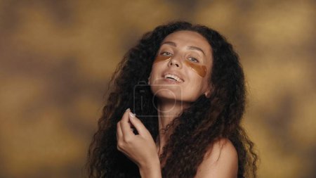 Photo for Longhaired curly brunette woman performs cosmetic procedures. Portrait of a seminude woman with golden patches under her eyes in the studio on a yellow background with highlights close up. The concept - Royalty Free Image