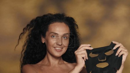 Téléchargez les photos : Une femme tient un masque de drap cosmétique noir. Séminude femme dans le studio sur un fond jaune avec des faits saillants. Le concept de beauté, de cosmétologie, de soins - en image libre de droit