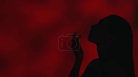 Foto de Primer plano de perfil lateral de una joven silueta femenina mirando hacia otro lado y levantando la mano hacia sus labios con gracia. Fondo rojo oscuro. Publicidad de ocio o producto. - Imagen libre de derechos