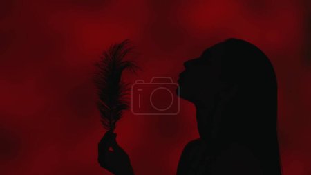 Foto de Vídeo de cerca del perfil lateral de una atractiva silueta de mujer joven que sopla en la pluma de su mano. Fondo rojo oscuro. Publicidad de ocio o producto. - Imagen libre de derechos