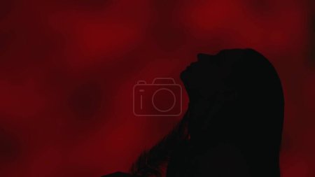 Foto de Primer plano de perfil lateral de una silueta de mujer joven graciosamente inclinada hacia atrás con la cabeza hacia arriba. Fondo rojo oscuro. Publicidad de ocio o producto. - Imagen libre de derechos