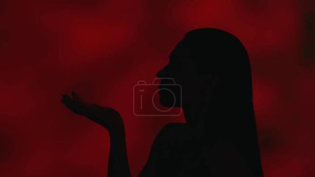 Téléchargez les photos : Gros plan profil vidéo d'une jeune femme silhouette soufflant une plume loin de sa paume. Fond rouge foncé. Loisirs ou publicité produit. - en image libre de droit