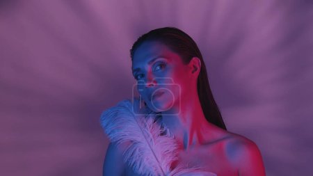 Photo for Young seminaked woman in close up view looking at the camera and covering herself with two big white feathers. Contrasting pink and blue color scheme, background covered in soft shadows. - Royalty Free Image