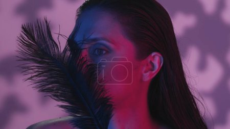 Photo for Close up side profile shot of a attractive young woman with a feather in front of her face. The woman seems relaxed. Neon pink and blue background, soft shadows. Leisure or product advertisement. - Royalty Free Image