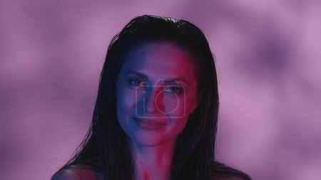 Photo for Close up shot of a young womans face. She is confidently looking at the camera in joyful, flirting and alluring manner, neon pink and blue color scheme. Leisure or product advertisement. - Royalty Free Image