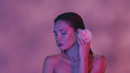 Photo for Close up shot of an attractive young seminaked woman. She is looking to the side and is holding a pink rose flower in her hand, next to her ear. Pink color scheme. Creative content. - Royalty Free Image