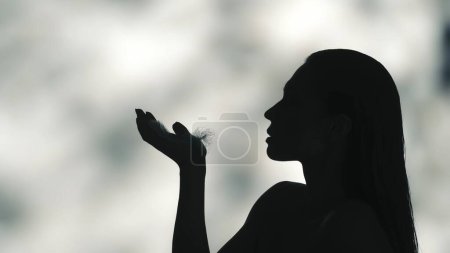 Téléchargez les photos : Gros plan profil latéral vidéo d'une jeune femme silhouette noire. Elle soufflait une petite plume sur sa paume. Fond vert clair ombragé. Loisirs ou publicité produit. - en image libre de droit