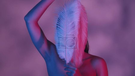 Photo for Close up shot of a seminude young woman in front of the camera, covering her face with two white feathers. Shadowed background. Pink and blue color scheme. Leisure or product advertisement. - Royalty Free Image