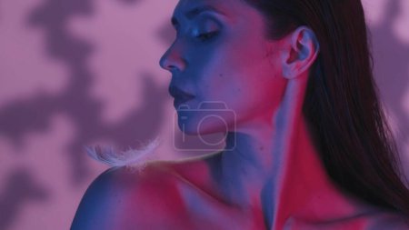 Photo for Close up side profile shot of a young woman with a feather on her shoulder. The woman is looking at it peacefully. Neon pink and blue background. Leisure or product advertisement. - Royalty Free Image