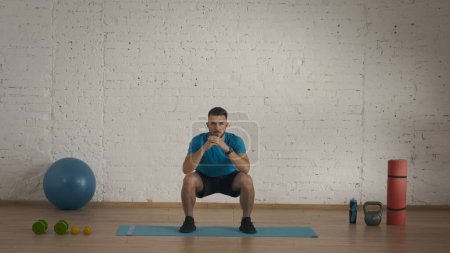 Photo for Sport indoors. Athletic man fitness coach doing exercises at the home studio for online classes. Man in sportswear doing standart squats. Healthcare concept. - Royalty Free Image